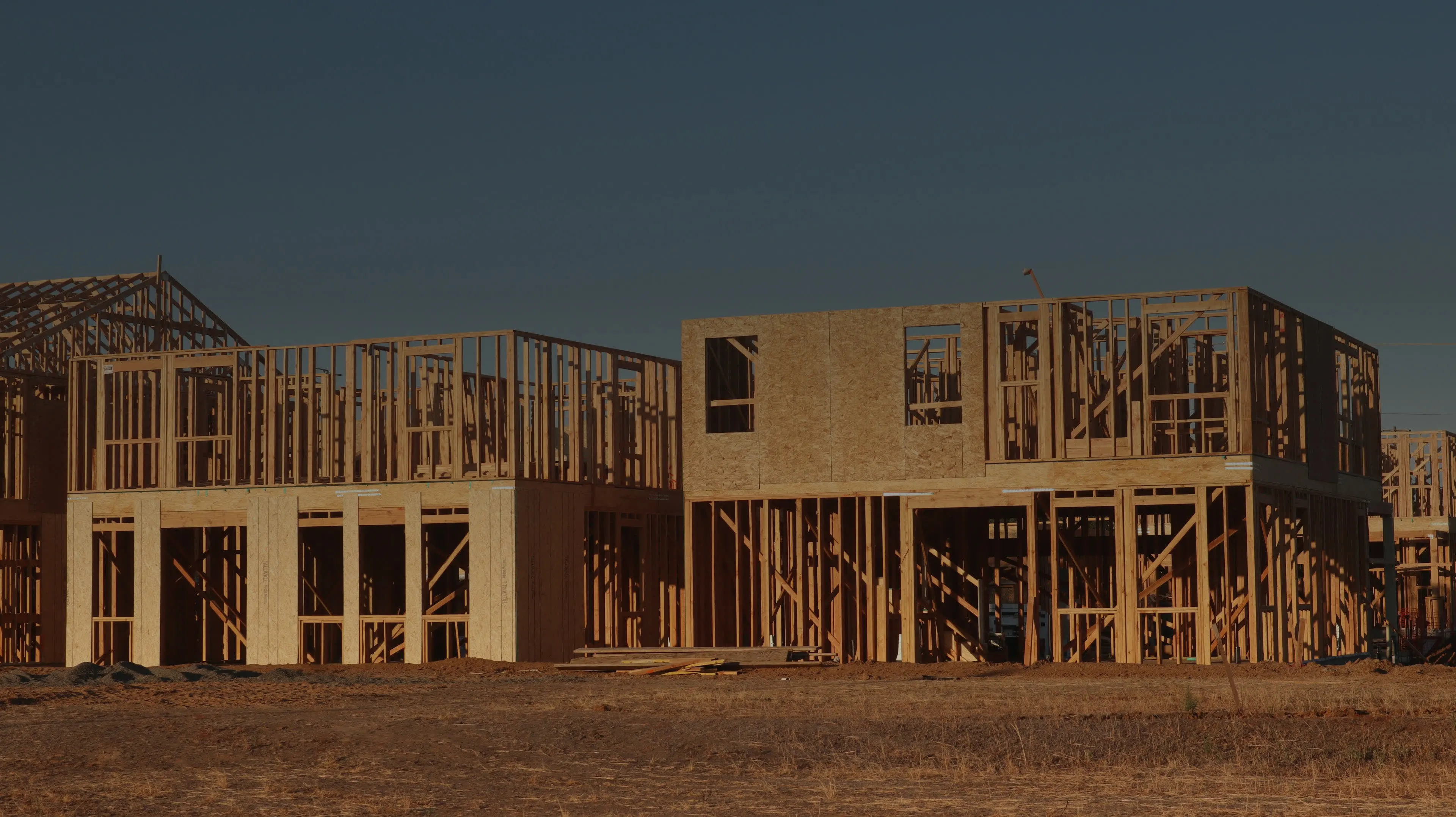 Houses at the framing stage in late afternoon light, open studs against a clear sky